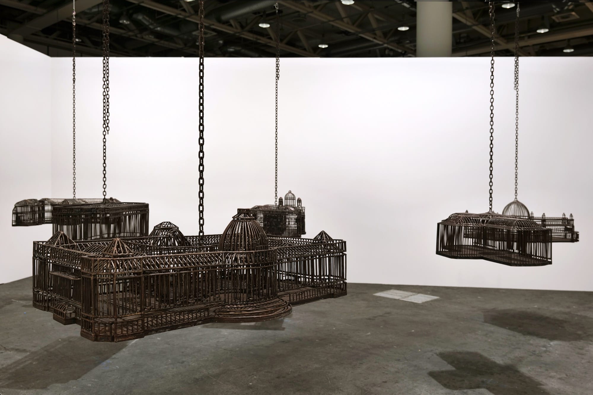 detail of an installation in a white-walled gallery space of sculptures suspended from the ceiling by chains, each one modeled after a colonial, institutional building