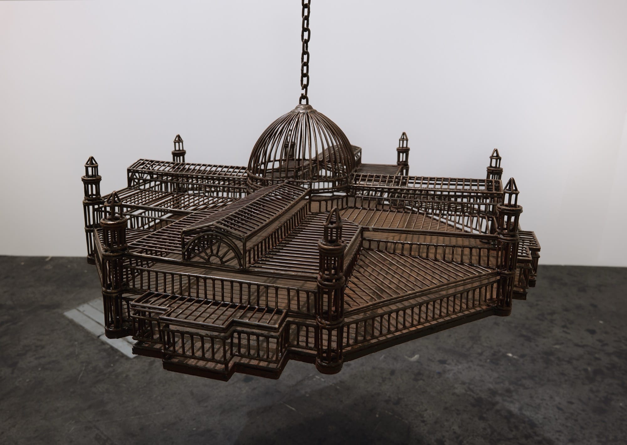 detail of an installation in a white-walled gallery space of sculptures suspended from the ceiling by chains, each one modeled after a colonial, institutional building