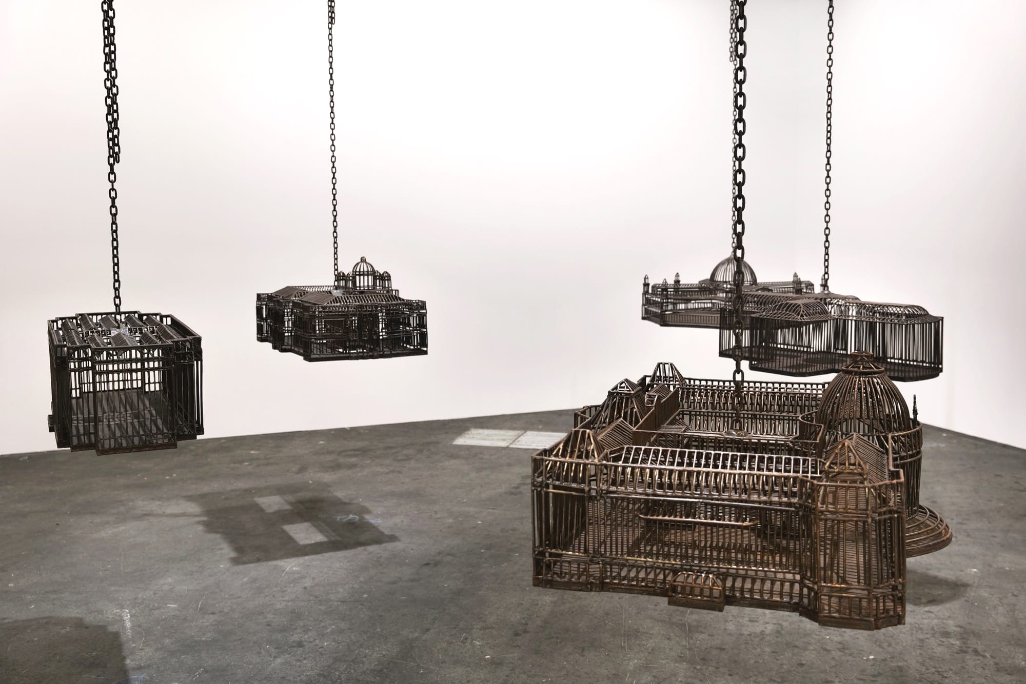 detail of an installation in a white-walled gallery space of sculptures suspended from the ceiling by chains, each one modeled after a colonial, institutional building