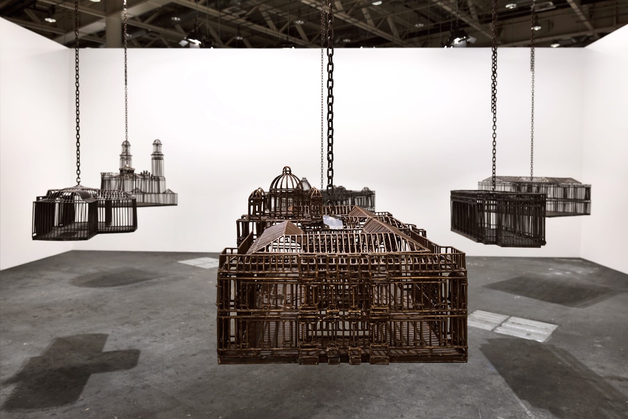 detail of an installation in a white-walled gallery space of sculptures suspended from the ceiling by chains, each one modeled after a colonial, institutional building