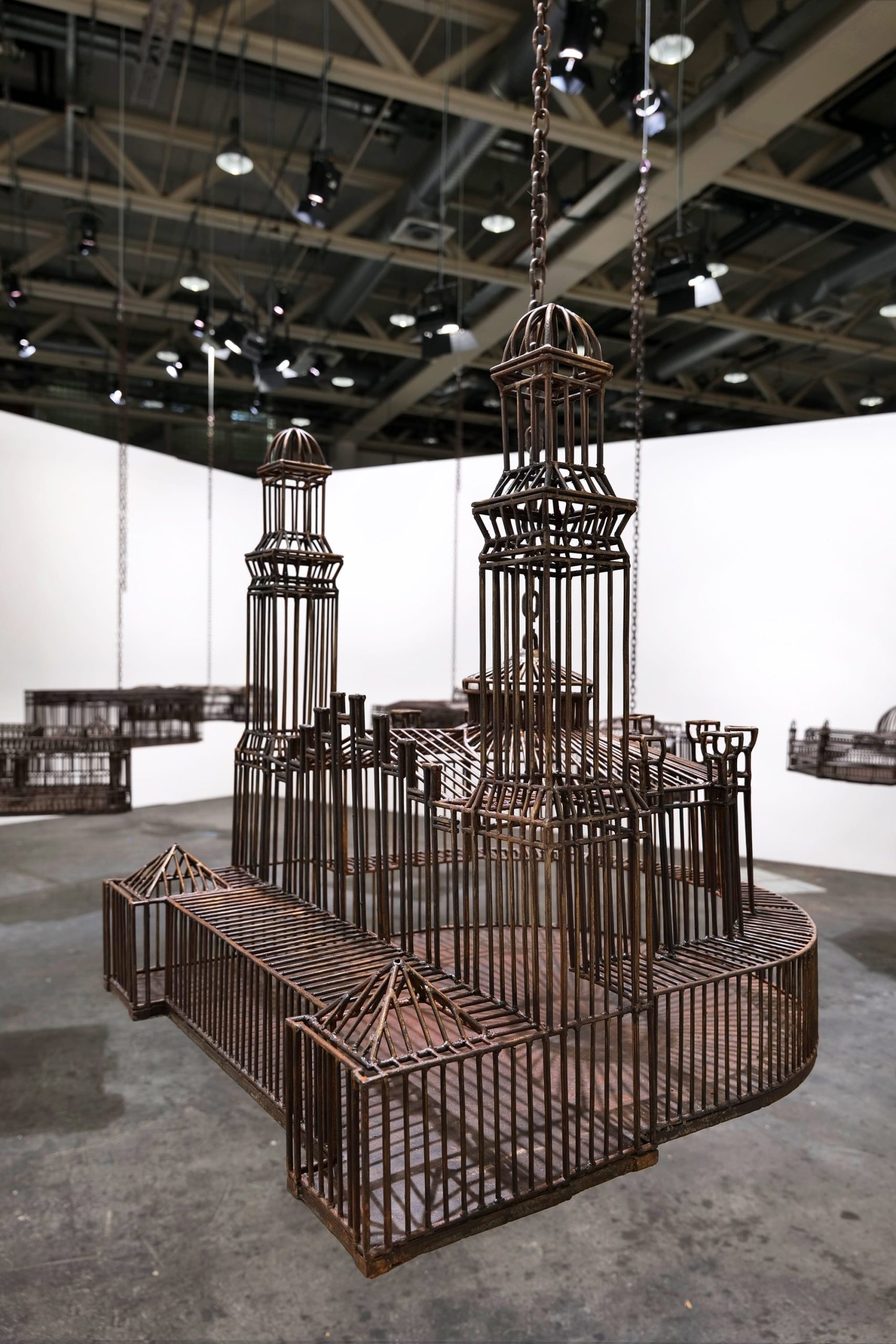detail of an installation in a white-walled gallery space of sculptures suspended from the ceiling by chains, each one modeled after a colonial, institutional building
