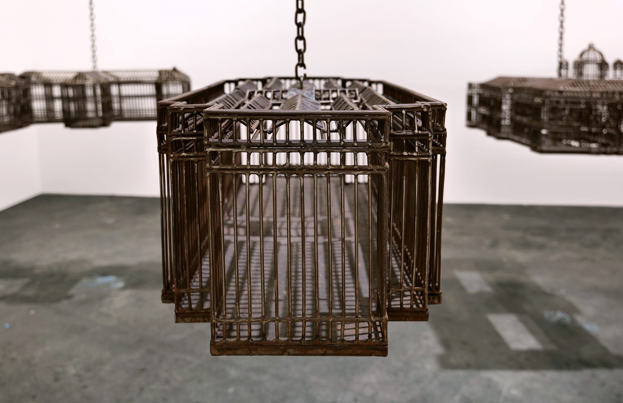 detail of an installation in a white-walled gallery space of sculptures suspended from the ceiling by chains, each one modeled after a colonial, institutional building
