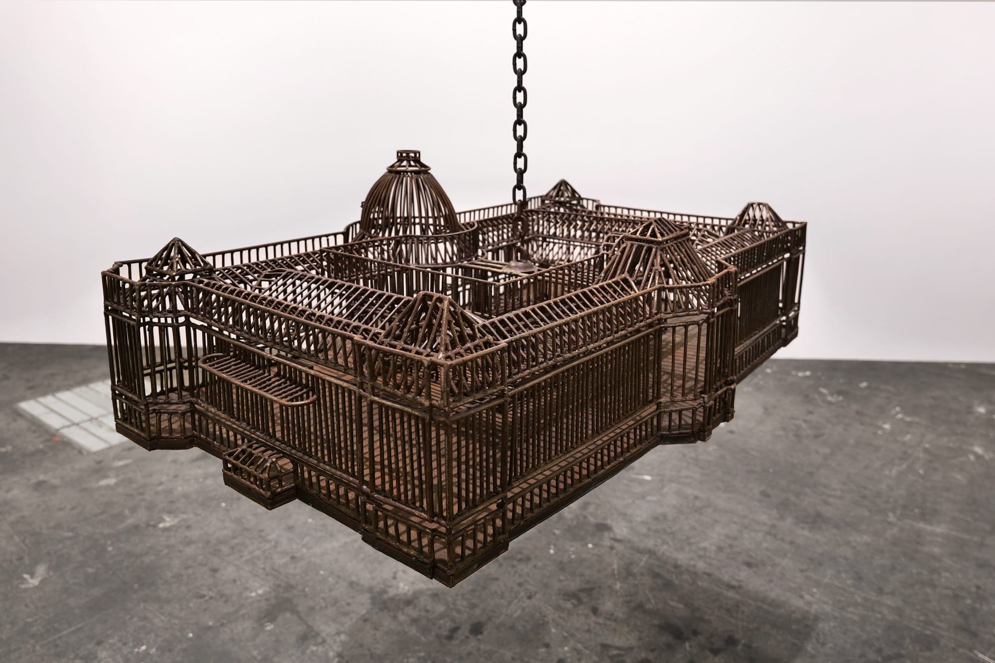 detail of an installation in a white-walled gallery space of sculptures suspended from the ceiling by chains, each one modeled after a colonial, institutional building
