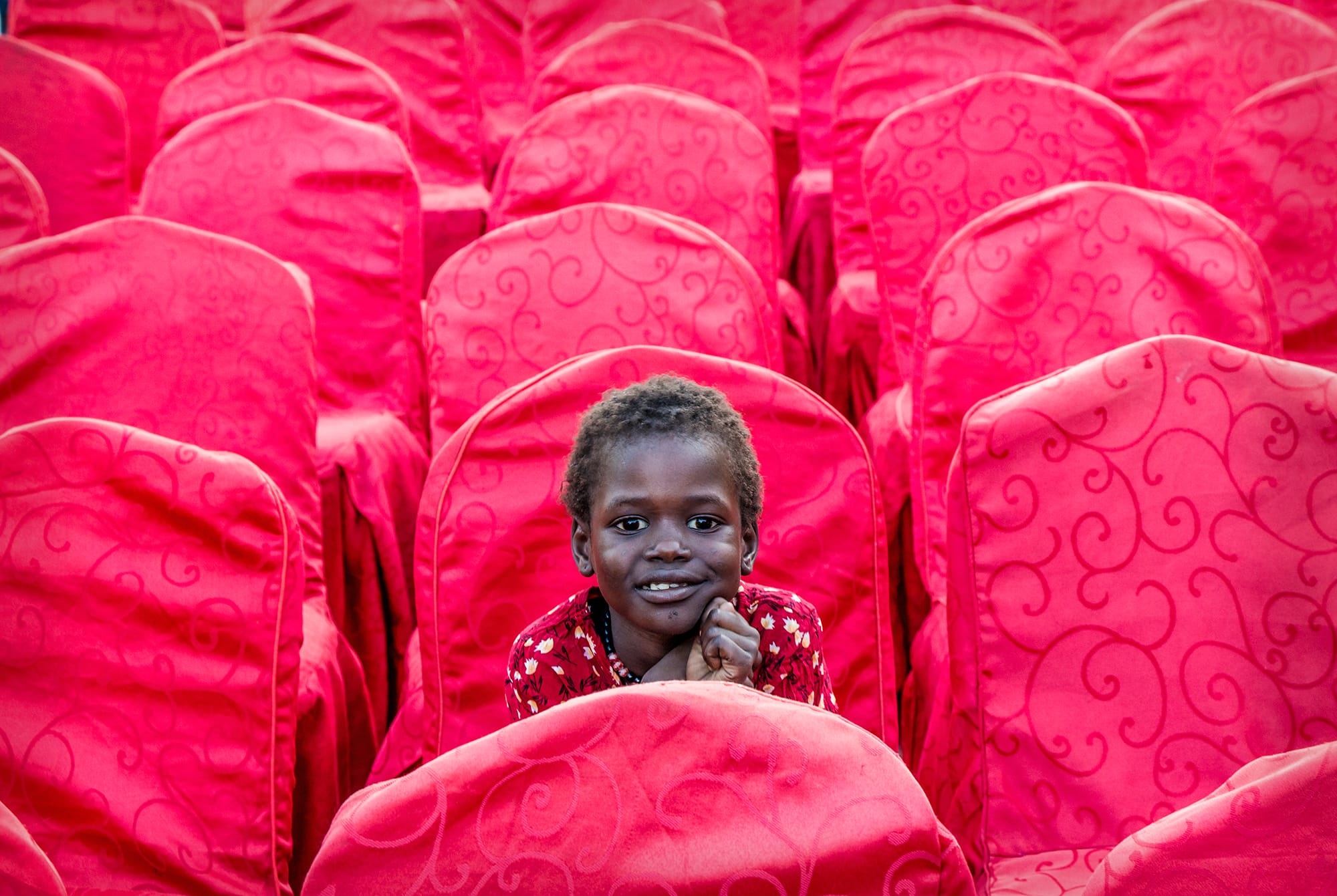 a photo by Altayeb Morhal of a young child seated alone amid a seat of bright pink-covered chairs