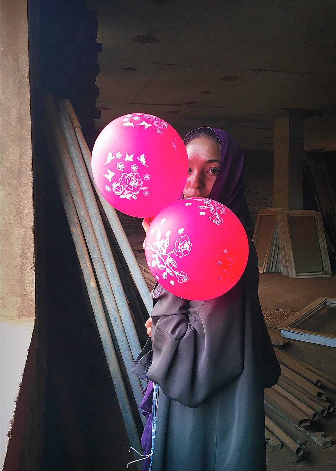 a photograph by Shaima Merghani of a young Sudanese woman standing near a window and holding two pink balloons